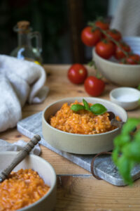 Tomatenrisotto in einer beigen Schale auf einem Marmorbrettchen. ImHintergrund sieht man frische Tomaten und im Vordergrung etwas unscharf Basilikum.