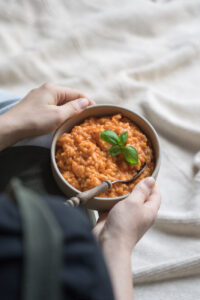 Tomatenrisotto in einer beigen Schale , von zwei Händen über einer hellen Decke gehalten.