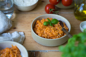 Tomatenrisotto in einer beigen Schale auf einem Marmorbrettchen. ImHintergrund sieht man frische Tomaten und im Vordergrung etwas unscharf Basilikum.