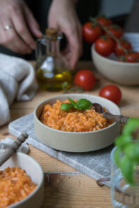 Tomatenrisotto in einer beigen Schale auf einem Marmorbrettchen. ImHintergrund sieht man frische Tomaten und im Vordergrung etwas unscharf Basilikum.