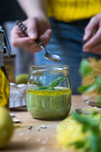 Ein Schraubglas mit Zitronen-Pesto, im Hintergrund steht ein Vorratsglas mit Pasta, eine Hand mit einem kleinen Löffel, der etwas Pesto entnehmen möchte, vorne erkennt man eine Zitrone und eine Ölflasche am Rand.