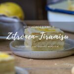 Ein Stück Zitronen-Tiramisu auf einem kleinen Teller, auf einem Holztisch Im Hintergrund sieht man eine Schale mit Tiramisu.