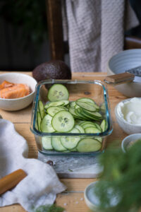 Eine Glasschale mit gehobelten Gurkenscheiben, kleine Schalen mit den weiteren Zutaten für einen Gurkensalat mit Lachs stehen darum.