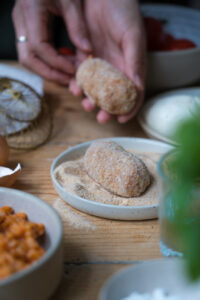 Zubereitung von Suppli, italienische Reiskroketten. Eine der rohen Kroketten liegt auf einem kleinen Teller mit paniermehl, im Hintergrund wird eine weitere gehalten. Die Tischplatte ist aus Holz. Man sieht unscharf Basilikum und eine Schale mit Risotto im Vordergrund.