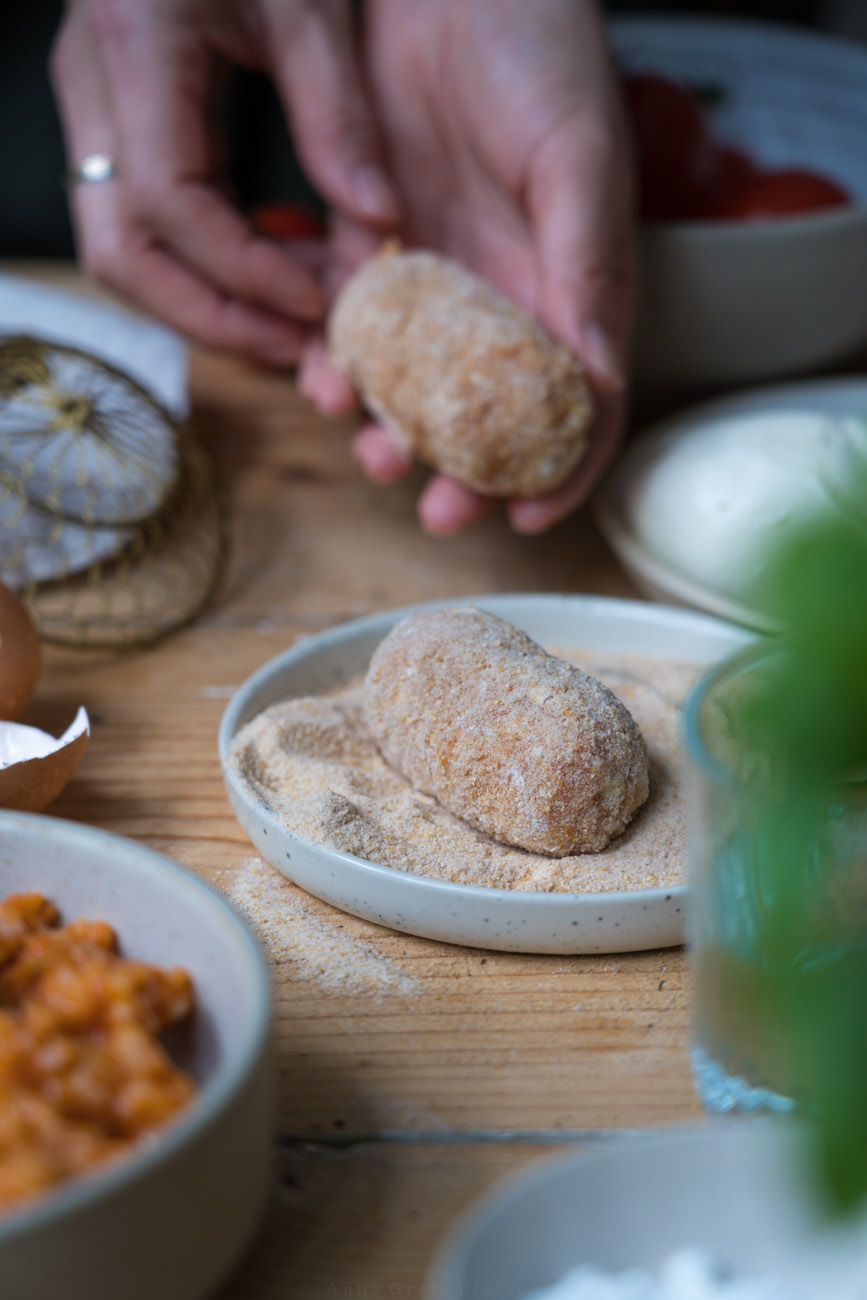 Zubereitung von Suppli, italienische Reiskroketten. Eine der rohen Kroketten liegt auf einem kleinen Teller mit paniermehl, im Hintergrund wird eine weitere gehalten. Die Tischplatte ist aus Holz. Man sieht unscharf Basilikum und eine Schale mit Risotto im Vordergrund.