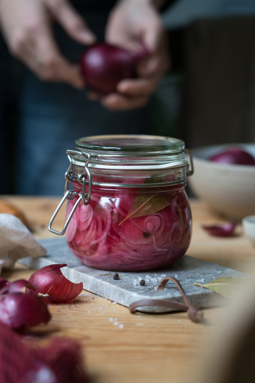 Eingelegte pinke Zwiebeln in einem Einmachglas. Der Untergrund ist ein Holztisch und ein Marmorbrettchen.