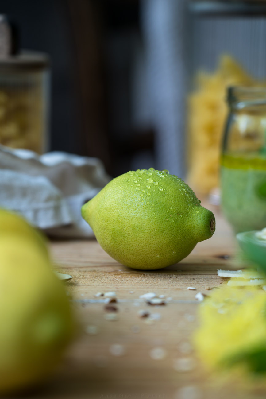 Eine frische Zitrone mit Wassertropfen liegt auf einem Holztisch, man sieht weitere Zitronen im Vordergrund, Vorratsgläser mit Nudeln im Hintergrund.