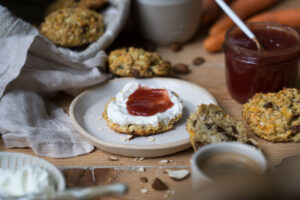 Ein Holztisch mit handgemachter, heller Keramit, eine Frühstücksszene, ein Brotkorb aus beigem Stoff, in der Mitte liegt ein Brötchen aus Haferflocken und Rosinen mit Quark und Erdbeermarmelade.