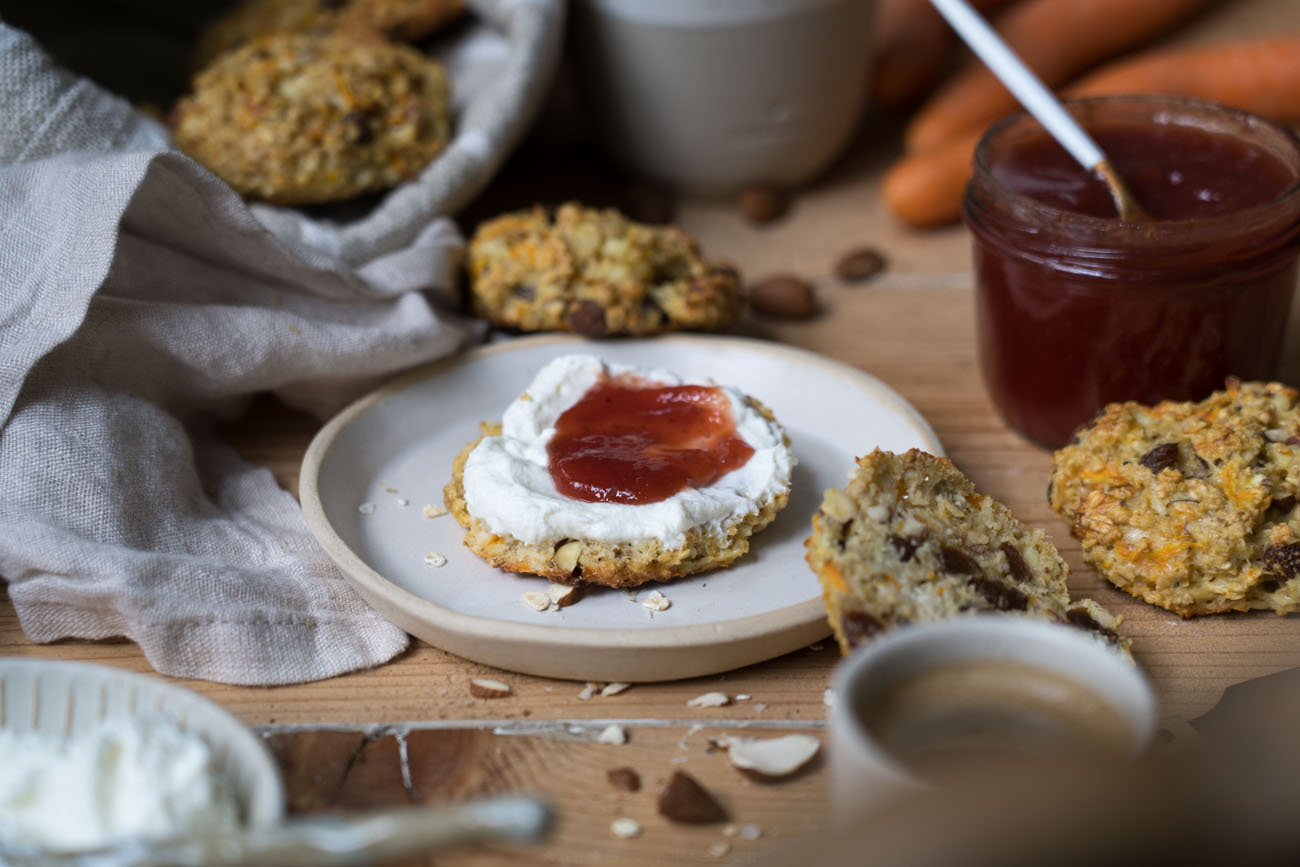 Ein Holztisch mit handgemachter, heller Keramit, eine Frühstücksszene, ein Brotkorb aus beigem Stoff, in der Mitte liegt ein Brötchen aus Haferflocken und Rosinen mit Quark und Erdbeermarmelade.