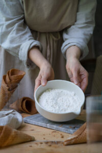 glutenfreies Mehl in einer hellen Schüssel, gehalten von zwei Händen, auf einer kleinen Marmorplatte, die auf einem Holztisch steht