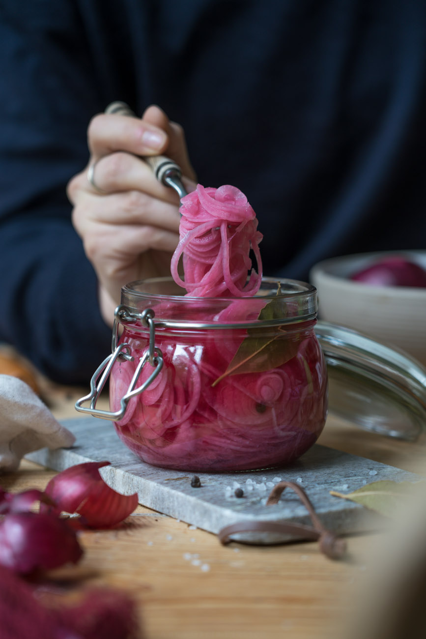 Eingelegte pinke Zwiebeln in einem Einmachglas. eine Gabel voll wird aus dem Glas genommen. Der Untergrund ist ein Holztisch und ein Marmorbrettchen.