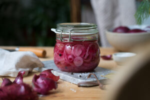 Eingelegte pinke Zwiebeln in einem Einmachglas. Der Untergrund ist ein Holztisch und ein Marmorbrettchen.