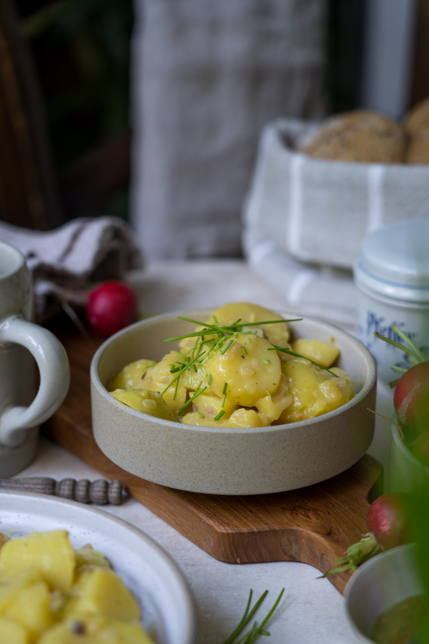Ein gedeckter Tisch. In der Mitte steht eine Schale mit baxerischem Kartoffelsalat, rundherum stehen ein Korb mit Brötchen, Radieschen, ein Bierkrug.