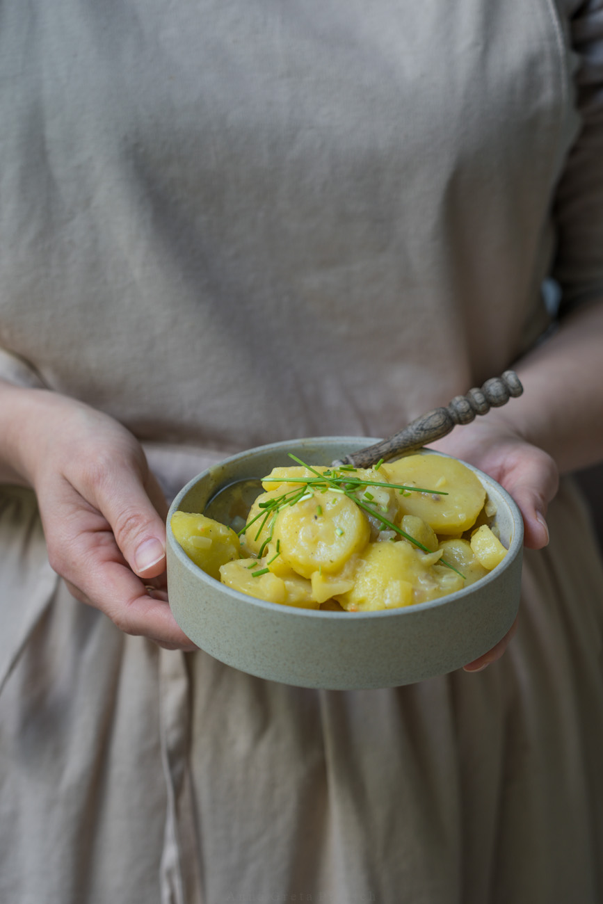 Eine Porzellanschale mit bayerischem Kartoffelsalat, gehalten von einer Person mit einer beigen Kochschürze.