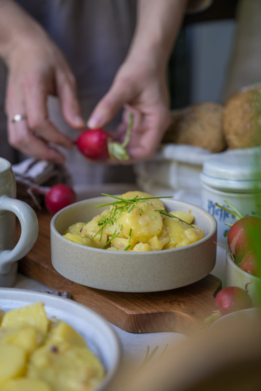 Ein gedeckter Tisch. In der Mitte steht eine Schale mit baxerischem Kartoffelsalat, rundherum stehen ein Korb mit Brötchen, Radieschen, ein Bierkrug.
