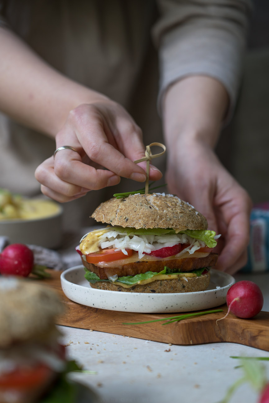 Ein Holzbrettchen mit einem selbstgemachten Leberkäseburger. Im Vorder- und Hintergrund sieht man einen gedeckten Tisch mit Kartoffelsalat, Brötchen und Radieschen. Eine Person steckt ein Spießchen in den Burger.