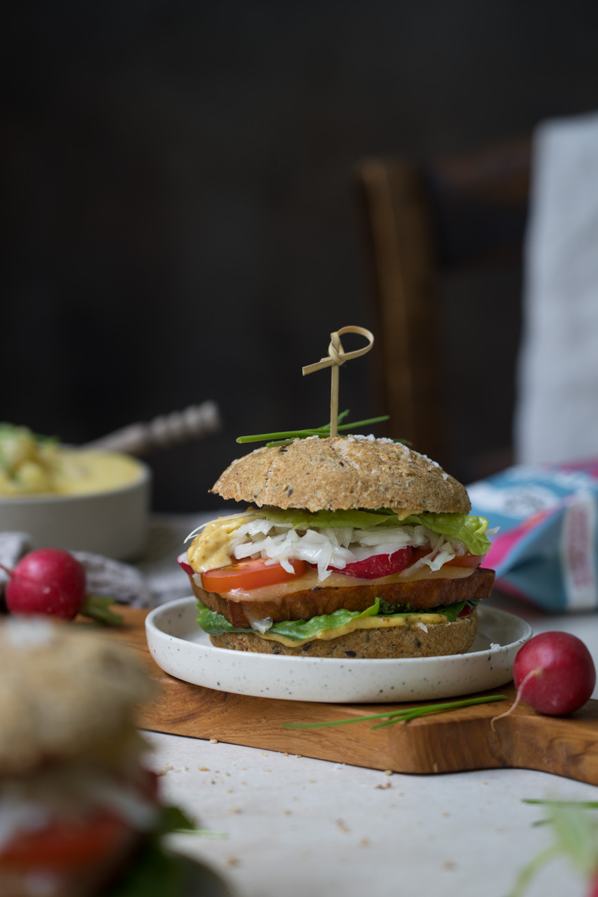Ein Holzbrettchen mit einem selbstgemachten Leberkäseburger. Im Vorder- und Hintergrund sieht man einen gedeckten Tisch mit Kartoffelsalat, Brötchen und Radieschen.