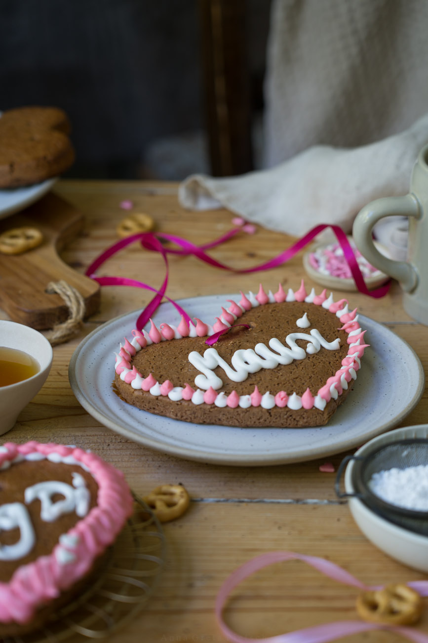 Ein Holztisch mit einem hellen Keramikteller, auf dem ein Lebkuchenherz mit der Aufschrift "Mausi" liegt.