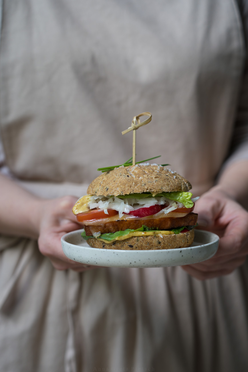 Ein Burger aus Laugenbrötchen mit Leberkäse, Soße, Salat und Dip. Wird gehalten von zwei Händen.