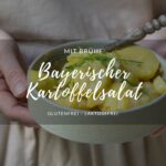 Ein Rezept für Bayerischen Kartoffelsalat. Eine Porzellanschale mit bayerischem Kartoffelsalat, gehalten von einer Person mit einer beigen Kochschürze.