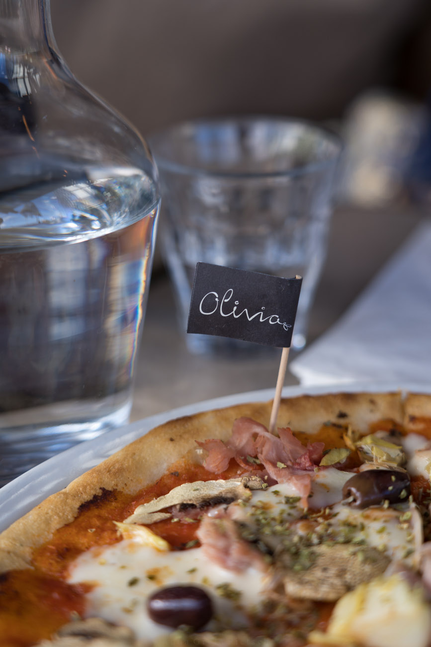 Ein Tisch in einem Restaurant mit einem Teller voller Pizza. In der Pizza steckt ein kleines Fähnchen mit der Aufschrift "Olivia" und auf der anderen Seite mit "glutenfri". Im Hintergrund steht eine Wasserkaraffe und ein Glas mit Wasser. Die Pizza ist belegt mit Champignons, Oliven, Schinken, Tomatensoße und Käse.