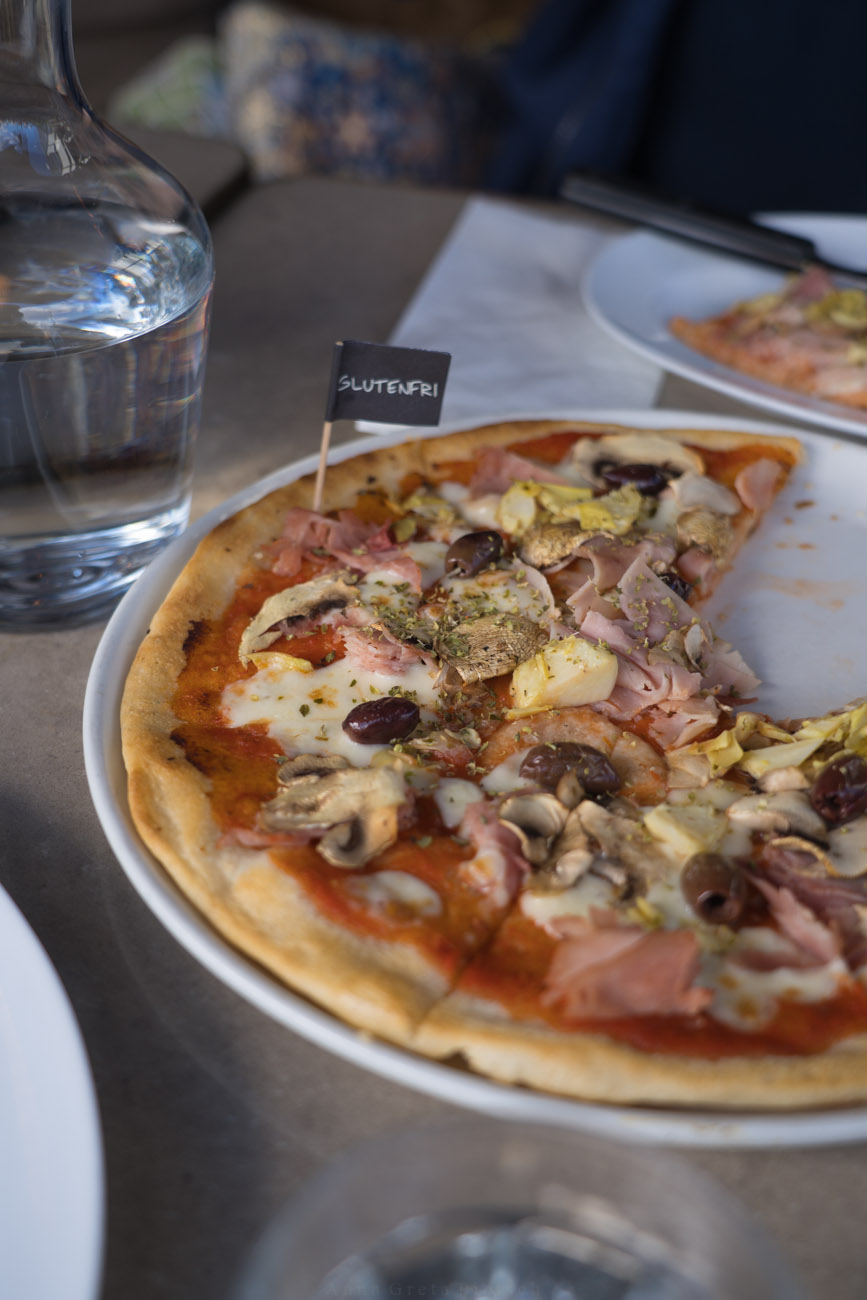Ein Tisch in einem Restaurant mit einem Teller voller Pizza. Im Hintergrund steht eine Wasserkaraffe. Die Pizza ist belegt mit Champignons, Oliven, Schinken, Tomatensoße und Käse.