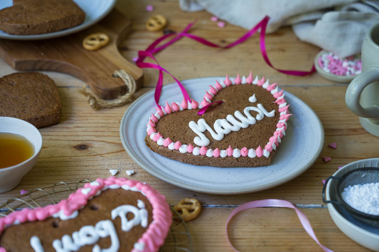 Ein Holztisch mit einem hellen Keramikteller, auf dem ein Lebkuchenherz mit der Aufschrift "Mausi" liegt.