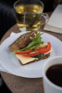 Ein weißer Teller mit zwei Scheiben Körnerbrot, belegt mit Käse, Salat udn Paprika. Vorne ist ein Anschnitt einer Kaffeetasse und im Hintergrund eine Teetasse zu sehen.