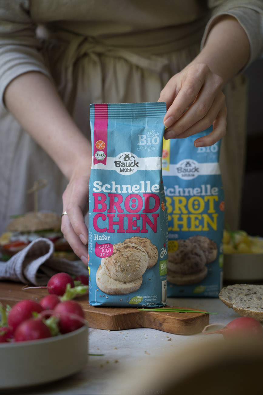 Eine Arbeitsfläche, in der Mitte steht eine Papierverpackung mit einer Backmischung für schnelle Brötchen, die von zwei Händen gehalten wird. Es luegen noch aufgeschnittene Brötchen und Radieschen auf der Arbeitsfläche.