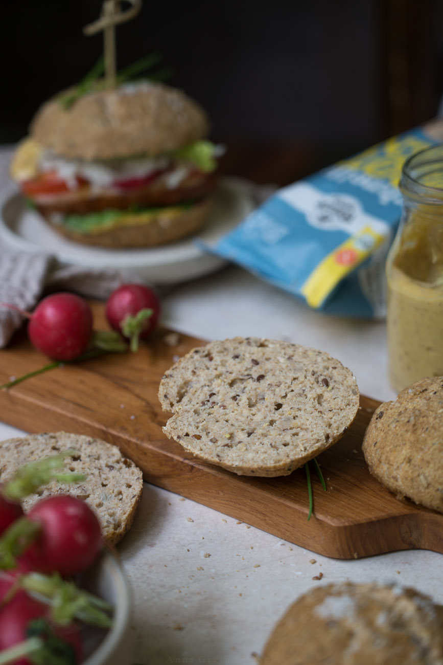 Ein Holzbrett auf einem hellen Untergrund mit einer aufgeschnittenen Brötchenhälfte. Drumherum liegen noch Radieschen, ein Burger und ein Glas mit einer Soße.