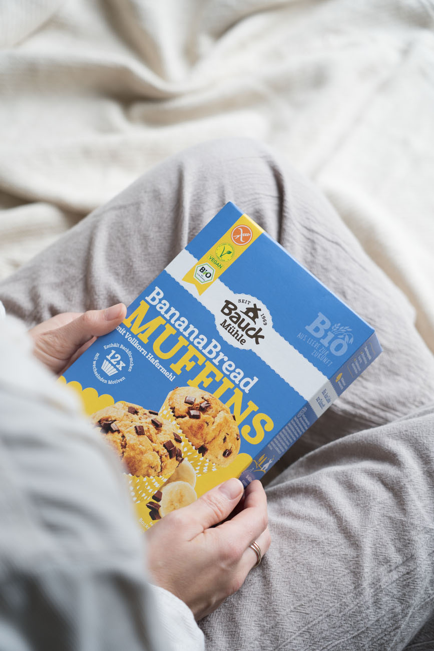 Eine Verpackung aus Karton von einer Backmischung für Banana Bread Muffins. Wird gehalten von zwei Händen, in einem Schoß liegend.