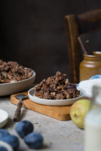 Eine Portion herbstlicher Crumble mit Zwetschgen, Birnen, Haferflocken und Kakao auf einem Tellerchen. Im Hintergrund sieht man die Schale mit Crumble und im Vordergrund ein Fläschchen mit Vanillesoße.