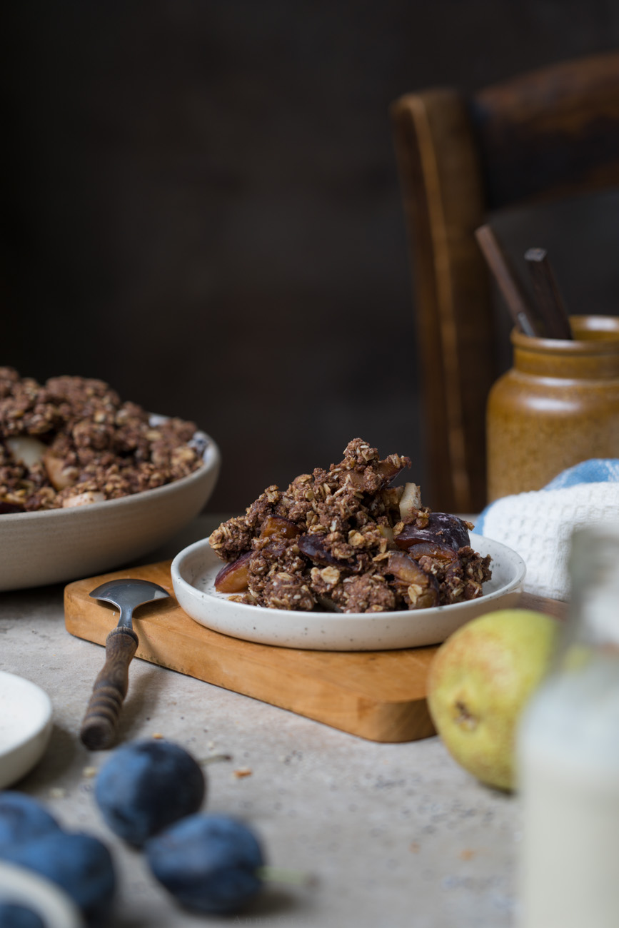 Eine Portion herbstlicher Crumble mit Zwetschgen, Birnen, Haferflocken und Kakao auf einem Tellerchen. Im Hintergrund sieht man die Schale mit Crumble und im Vordergrund ein Fläschchen mit Vanillesoße.