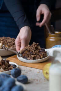 Eine Portion herbstlicher Crumble mit Zwetschgen, Birnen, Haferflocken und Kakao auf einem Tellerchen. Wird von einer Hand abgestellt. Im Hintergrund sieht man die Schale mit Crumble und im Vordergrund ein Fläschchen mit Vanillesoße.