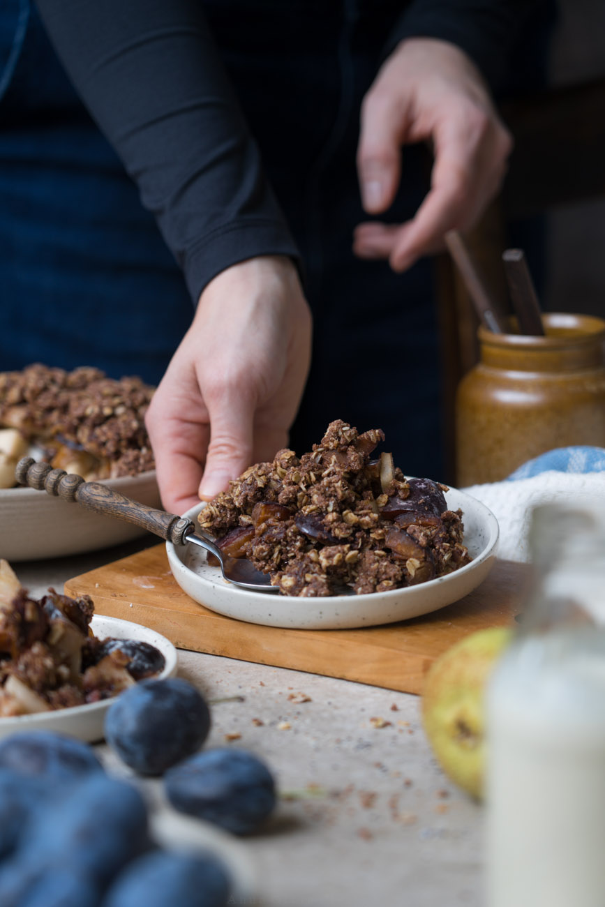 Eine Portion herbstlicher Crumble mit Zwetschgen, Birnen, Haferflocken und Kakao auf einem Tellerchen. Wird von einer Hand abgestellt. Im Hintergrund sieht man die Schale mit Crumble und im Vordergrund ein Fläschchen mit Vanillesoße.