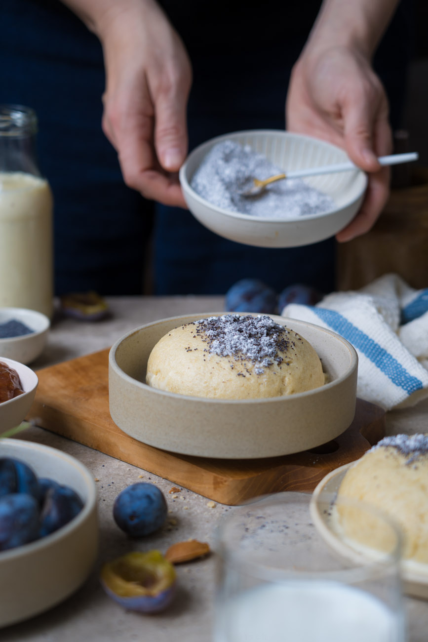 Eine Keramikschale mit einem Germknödel und Mohn-Zucker darauf. Im Vordergrund sieht man noch einen Knödel, Pflaumen in einer Schale. Im Hintergrund hält eine Person ein Schälchen mit Mohn-Zucker.