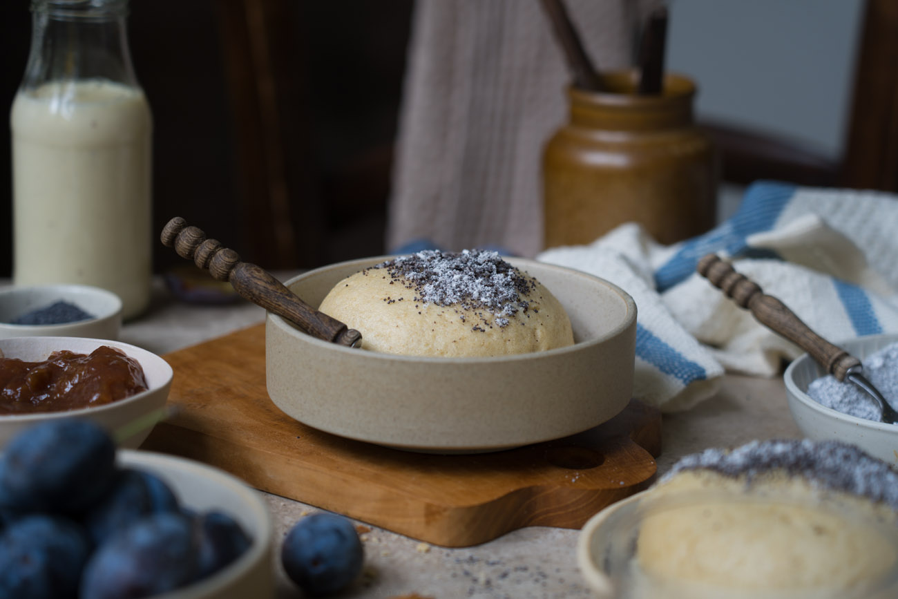 Eine Keramikschale mit einem Germknödel und Mohn-Zucker darauf. Im Vordergrund sieht man noch einen Knödel, Pflaumen in einer Schale.