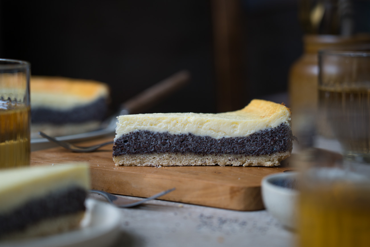 Ein Stück Mohn-Käsekuchen auf einem Holzbrett, im Hintergrund sieht man einen Teller mit Kuchen. Dazu stehen Gläser mit Tee auf dem Tisch.