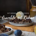Eine Portion herbstlicher Crumble mit Zwetschgen, Birnen, Haferflocken und Kakao auf einem Tellerchen. Im Hintergrund sieht man die Schale mit Crumble und im Vordergrund ein Fläschchen mit Vanillesoße.