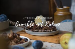 Eine Portion herbstlicher Crumble mit Zwetschgen, Birnen, Haferflocken und Kakao auf einem Tellerchen. Im Hintergrund sieht man die Schale mit Crumble und im Vordergrund ein Fläschchen mit Vanillesoße.