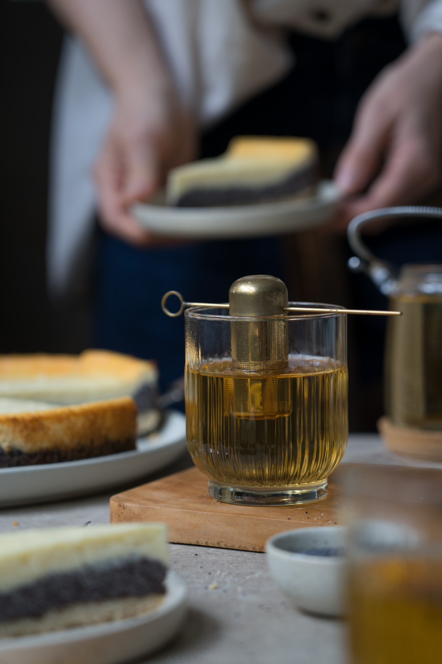 Ein Teeglas mit Teesieb und grünem Tee auf einem Holzbrettchen. Im Hintergrund sieht man eine PErson, die ein Stück Kuchen auf einem Teller hält und einen angeschnittenen Kuchen auf dem Tisch.