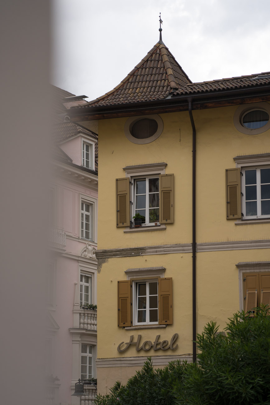 Ein gelbes Gebäude mit einem kleinen Spitzdach und Fensterläden aus Holz in Bozen.