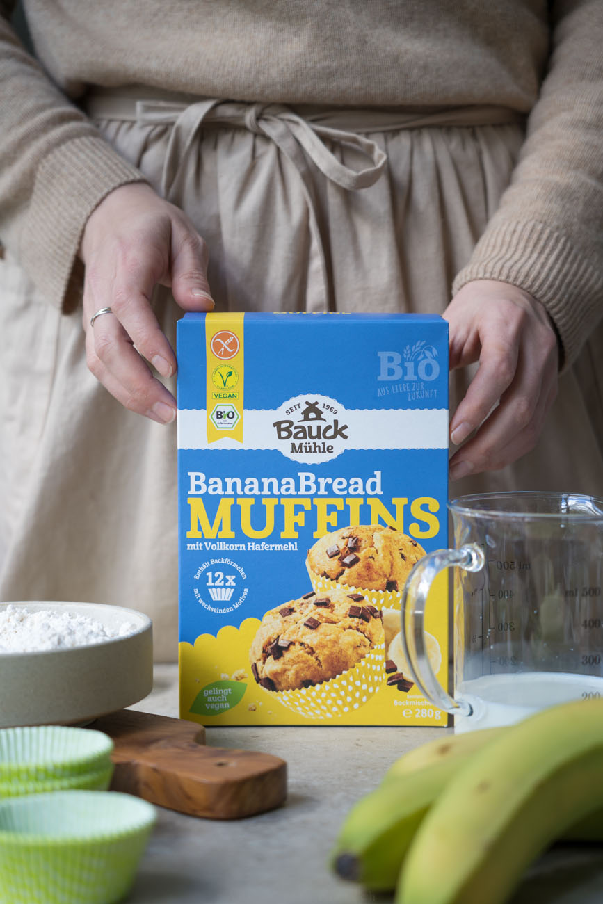 Eine Verpackung aus Karton von einer Backmischung für Banana Bread Muffins. Wird gehalten von zwei Händen, auf einem Küchentosch stehend.