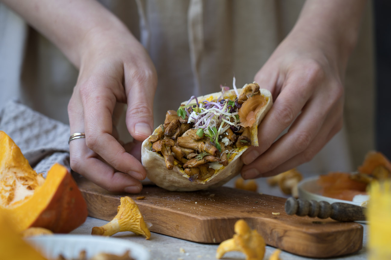 Gefüllte Pitataschen mit Pfifferlingen, gehalten von zwei Händen über einem Holzbrettchen. Im Hintergrung sieht man ein Glas mit orangefarbenem Dip.