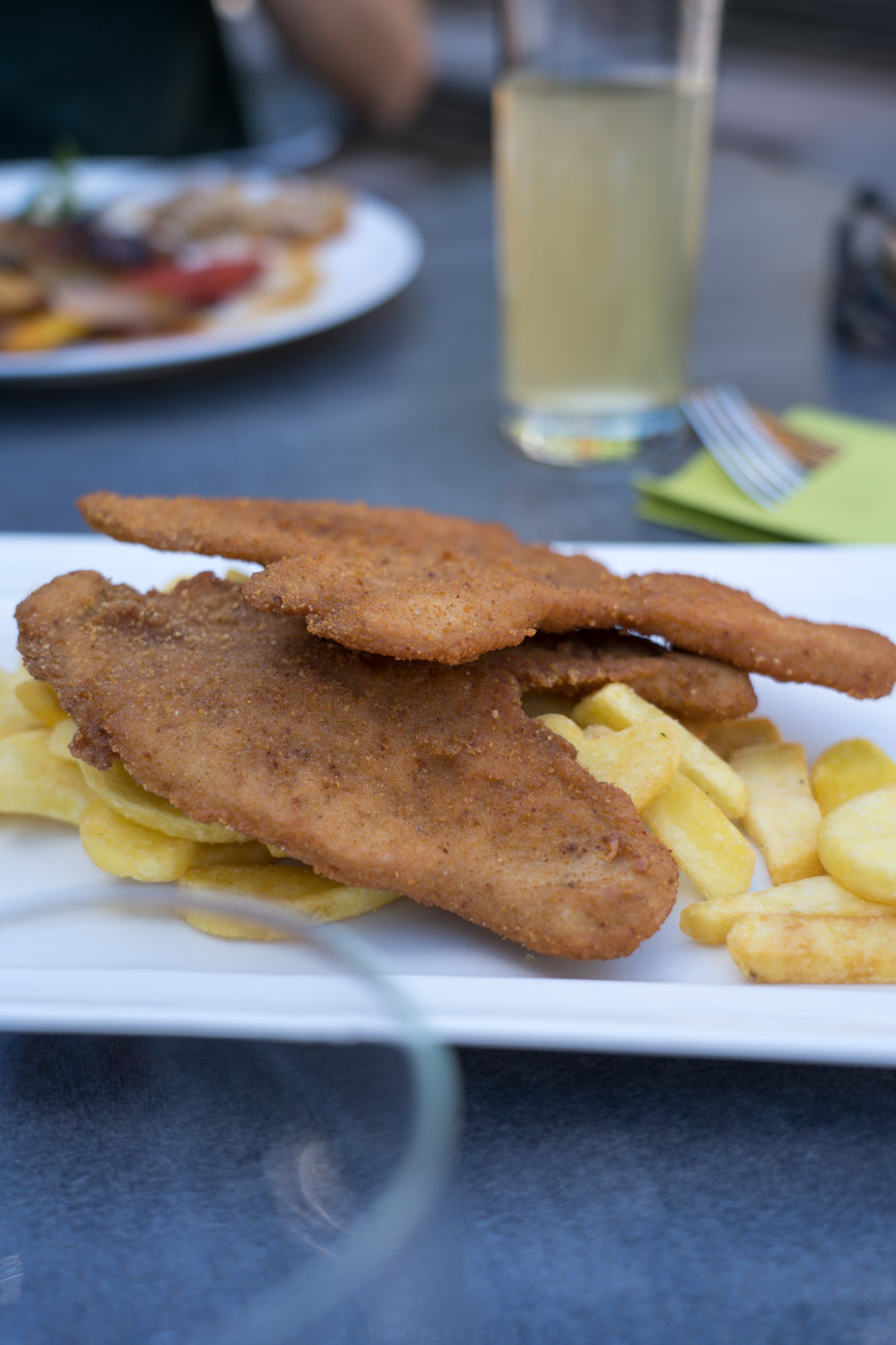 Ein weißer Teller mit Schnitzel und Pommes auf einem dunklen Tisch.