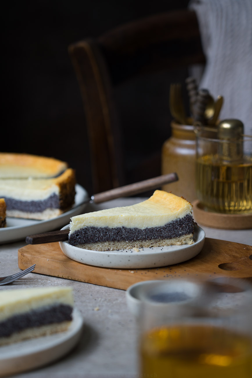 Ein Stück Mohn-Käsekuchen auf einem Holzbrett, im Hintergrund sieht man einen Teller mit Kuchen. Dazu stehen Gläser mit Tee auf dem Tisch.