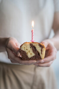 Ein Stück Marmorkuchen, gehalten von 2 Händen, mit einer rosafarbenen Geburtstagskerze, die brennt.