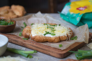 Ein glutenfreier Langos auf einem Holzbrett und einem Bogen Backpapier. Man sieht Besteck in einem Behälter im Hintergrund, geschnittene Frühlingszwiebel in einem Schälchen und eine Creme im Vordergrund.