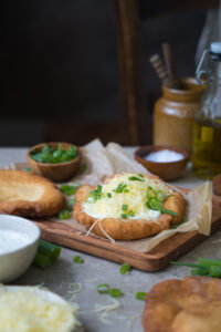 Ein glutenfreier Langos auf einem Holzbrett und einem Bogen Backpapier. Man sieht Besteck in einem Behälter im Hintergrund, geschnittene Frühlingszwiebel in einem Schälchen und eine Creme im Vordergrund.