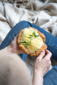 Ein glutenfreier Langos wird von zwei Händen gehalten. Das Foto wurde über die Schulter aufgenommen, so dass man noch eine Decke sieht, auf der die PErson sitzt und ihre Beine in einer Jeans.